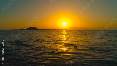 AERIAL Man sitting on his surfboard and outstretches his arms at stunning sunset
