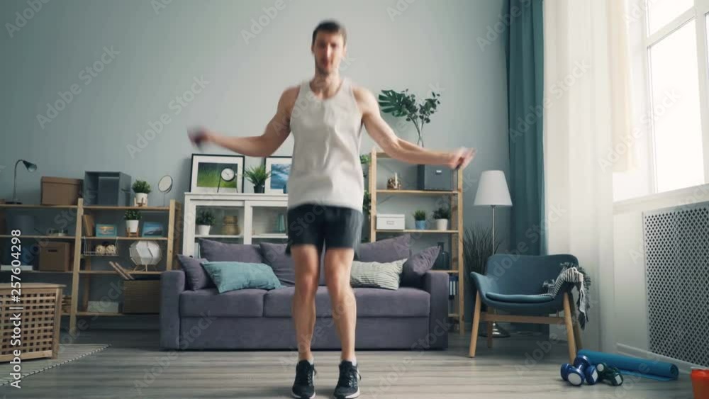 Sporty guy student is jumping rope in apartment exercising alone enjoying indoor sports wearing top, shorts and sneakers. Active lifestyle and modern youth concept. Sporty guy student is jumping rope in apartment exercising alone enjoying indoor sports wearing top, shorts and sneakers. Active lifestyle and modern youth concept.