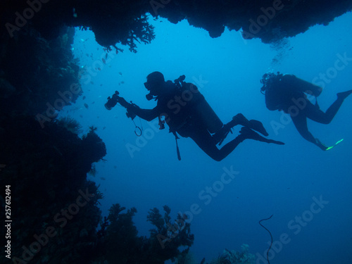Two divers are silhouetted through cave and coral © Alicia