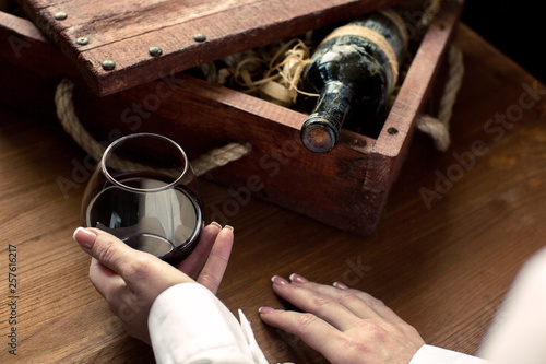 Human hand in white cuff with a glass of red wine. Old bottle in vintage box among wood shaving on wooden background, selected focus