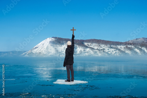 Man standing on ice floes floating in river in winter holding wooden cross with background of snow mountain photo