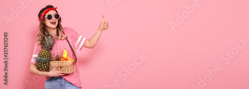 Beautiful young girl in pink t-shirt and glasses, holds a full straw bag of fruit on pink background. Shows finger class
