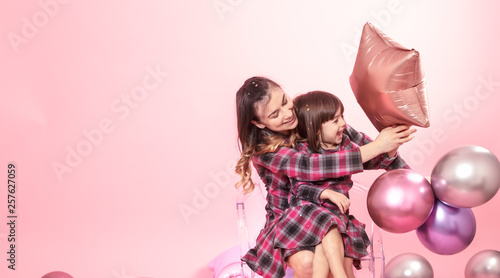 Funny mom and child sitting on a transparent stylish chairs pink background. Little girl and mother having fun with balloons and confetti
