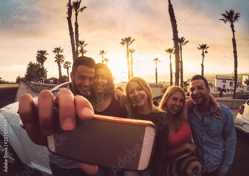 Happy friends standing in front of convertible car and taking selfie with mobile phone camera - Young millennial people having fun on roadtrip in tropical destination - Travel and technology concept