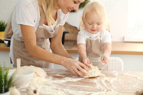 Little girl and her blonde mom in beige aprons  playing and laughing while kneading the dough in kitchen. Homemade pastry for bread, pizza or bake cookies. Family fun and cooking concept