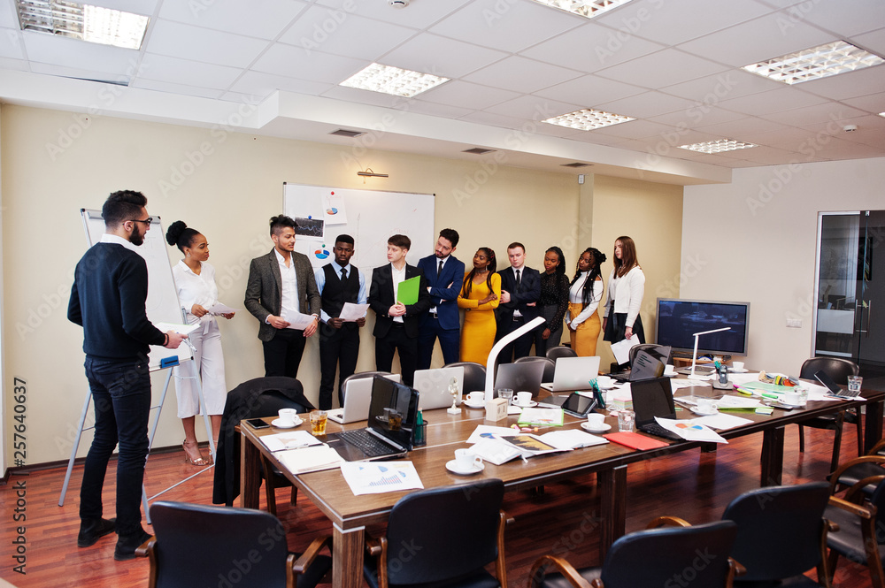 Mixed race business coach presenting report standing near whiteboard pointing on sales statistic shown on diagram and chart teach diverse company members gathered together in conference room. Mixed race business coach presenting report standing near whiteboard pointing on sales statistic shown on diagram and chart teach diverse company members gathered together in conference room.