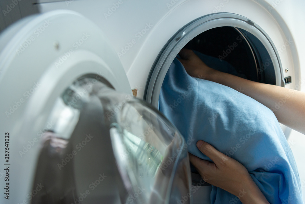 Close up of woman hands putting a blue bed sheet to wash in a washing machine Close up of woman hands putting a blue bed sheet to wash in a washing machine