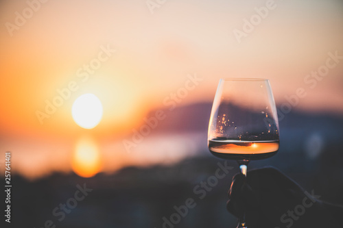 Man's hand holding glass of rose wine and with sea and beautiful sunset at background, close-up, horizontal composition. Summer evening relaxed mood concept