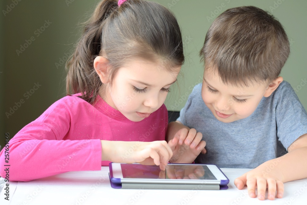 White caucasian toddler boy and beautiful girl smiling and playing together on tablet games. Portrait of a happiness little brother and sister. Happy Children play a computer game on the tablet. White caucasian toddler boy and beautiful girl smiling and playing together on tablet games. Portrait of a happiness little brother and sister. Happy Children play a computer game on the tablet.
