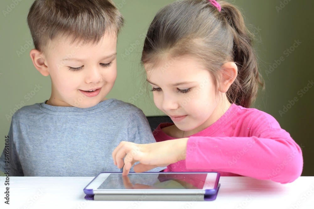 White caucasian toddler boy and beautiful girl smiling and playing together on tablet games. Portrait of a happiness little brother and sister. Happy Children play a computer game on the tablet. White caucasian toddler boy and beautiful girl smiling and playing together on tablet games. Portrait of a happiness little brother and sister. Happy Children play a computer game on the tablet.