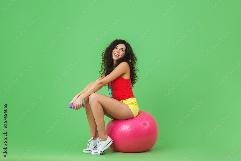 Image of delighted woman 20s wearing summer clothes holding dumbbells while sitting on fitness ball during aerobics Image of delighted woman 20s wearing summer clothes holding dumbbells while sitting on fitness ball during aerobics