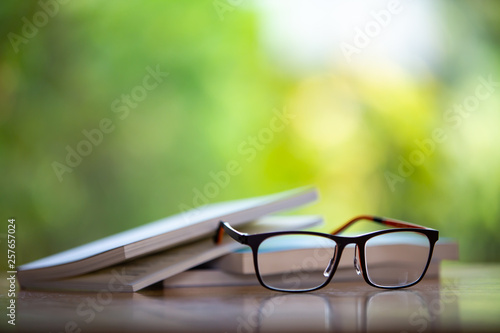 Black eyeglasses with books on wooden table, Bokeh garden background, Close up & Macro shot, Selective focus, Stationery concept