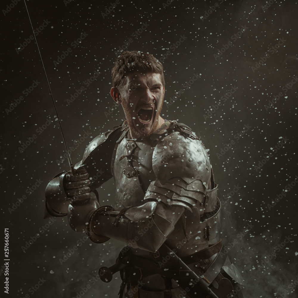Emotional portrait of a young man in knight armor and a sword against a dark background.