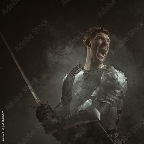 Fotografia, Obraz Emotional portrait of a young man in knight armor and a sword against a dark background