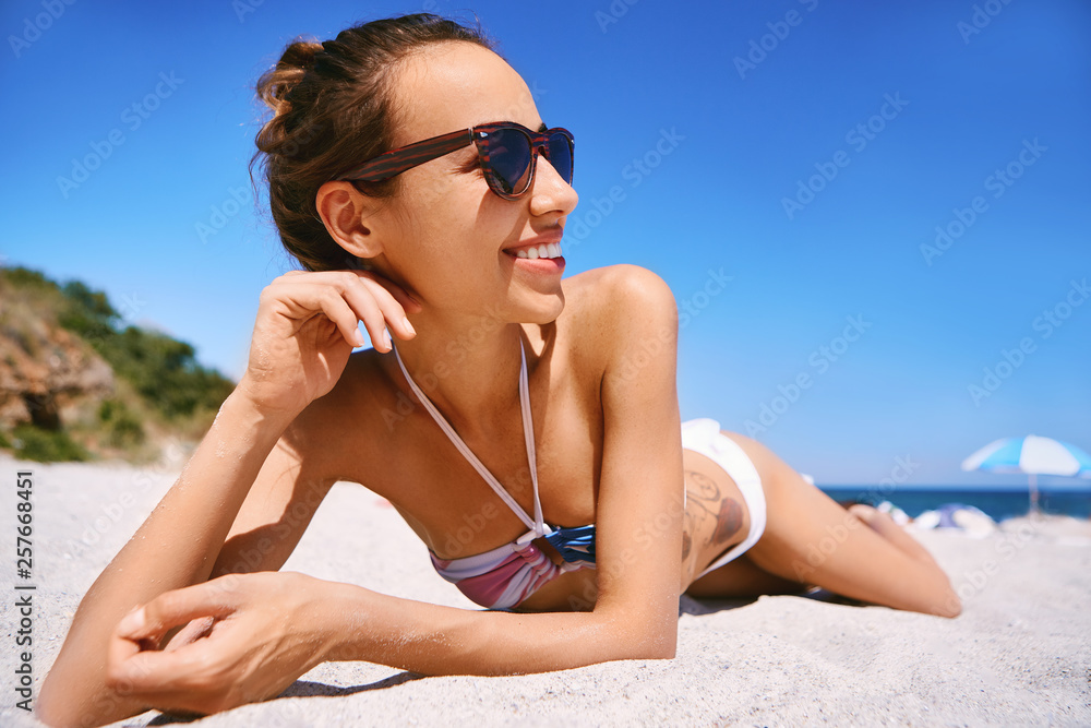 Charming smiling slim woman wearing in bikini is lying on white sand on the sea beach. Woman with sunglasses is sunbathing at heat sunny day at summer vacation Charming smiling slim woman wearing in bikini is lying on white sand on the sea beach. Woman with sunglasses is sunbathing at heat sunny day at summer vacation