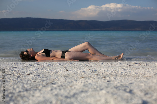 A young sexy female in a black swim suit is lying on a white sand of the turquoise crater lake Salda Golu, Turkey. Summer vacations. Dusk light. Copy space for the text. 