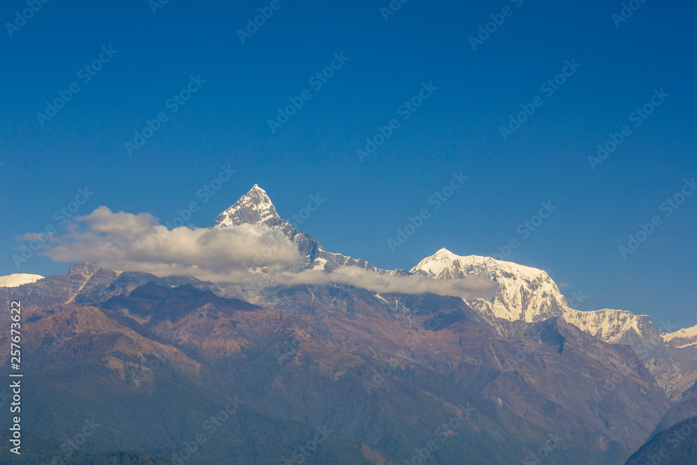 Obraz premium hillsides with green forests on the background of desert mountains and the snowy ridge of Annapurna with clouds under a clear blue sky. Machapuchare Himalayas Nepal