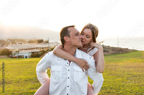 Romantic and emotional view of couple in white clothes when he carries her on the back. Beautiful landscape of sun above mountain during sunset and green grass lawn. Concept of honey moon love.
