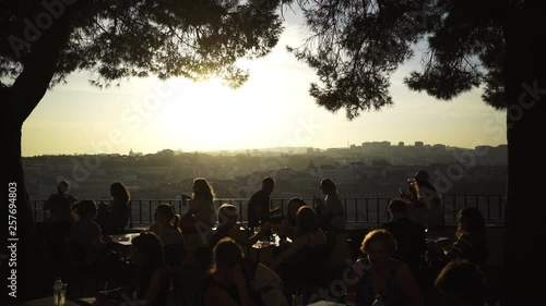 Silhouette people relaxing in street cafe on high top above city under trees and sky with sun in Lisbon