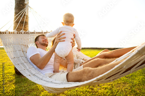 happy family on a tropical island at sunset lie in a hammock and play with their son