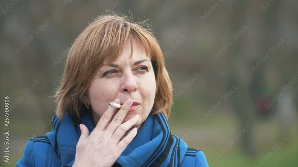 Confident blonde woman standing and smoking a cigarette in a park in slow motion Confident blonde woman standing and smoking a cigarette in a park in slow motion