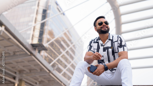 Young bearded Indian man with sunglasses sitting in the city outdoors