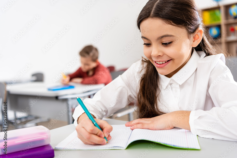 Smiling schoolgirl writing in notebook during lesson in classroom Smiling schoolgirl writing in notebook during lesson in classroom