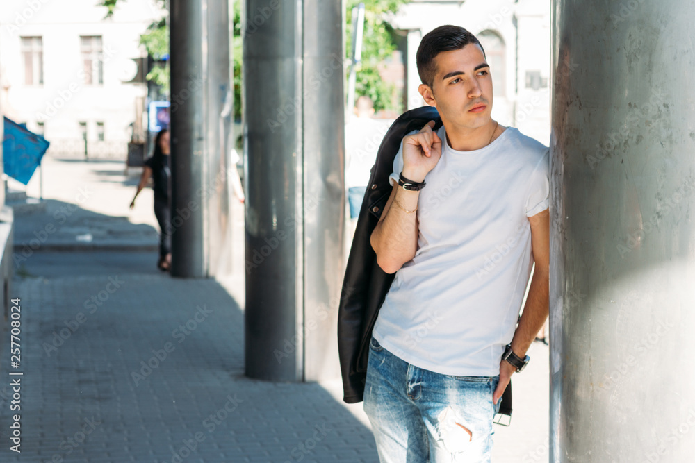 young, handsome guy in a white T-shirt, black, leather jacket and blue, torn jeans is on the street, model, city walk, live style. in white sneakers