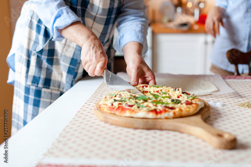 Woman cuts hot pizza on wooden board in cozy home kitchen. Cooking process of italian family dinner at home. Lifestyle moment. Close up of female hands with knife.