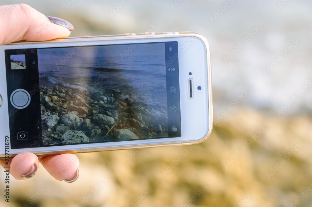 Woman hand with manicure holding white mobile phone with wave and sea in background.