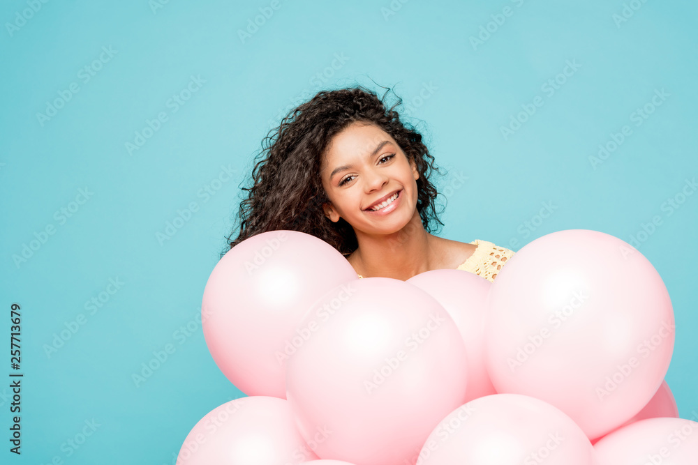 happy curly african american girl near pink air balloons isolated on blue happy curly african american girl near pink air balloons isolated on blue