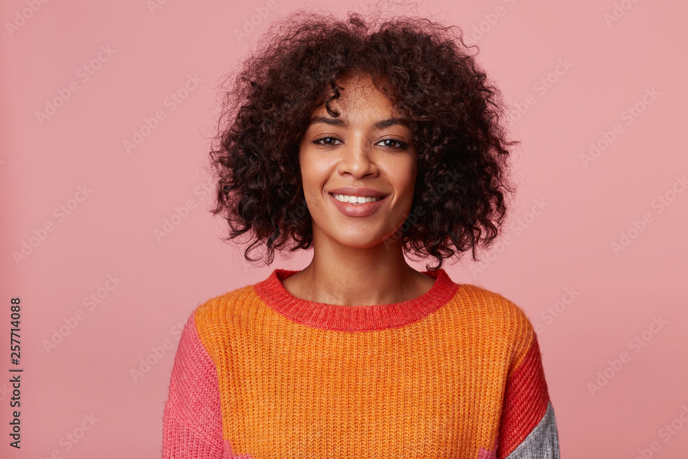Close up portrait of positive optimistic charming african american girl with afro hairstyle looks with pleasure, with friendly smile, wearing colorful longsleeve, isolated on pink background Close up portrait of positive optimistic charming african american girl with afro hairstyle looks with pleasure, with friendly smile, wearing colorful longsleeve, isolated on pink background