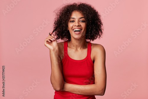 Attractive joyful african american girl with afro hairstyle looking swell wonderful plays with stand of curly dark hair  laughing  wearing red singlet  isolated on pink background