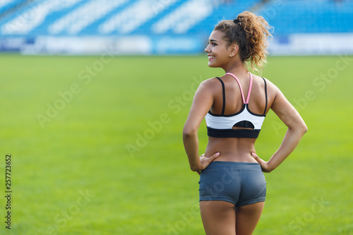 Young girl with curly hair and bronze tan in trendy sportswear standing back to the camera