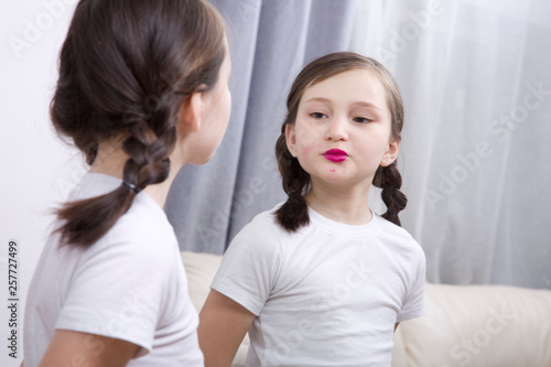 A little girl looks in the mirror and paints her lips with bright lipstick.