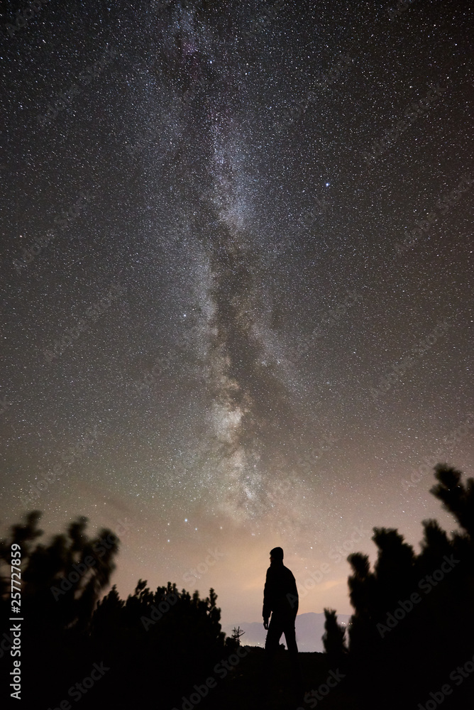 Silhouette of male hiker having a rest at summer night on the top of the mountain. Back view of man traveller enjoying view of night sky full of stars and Milky way.