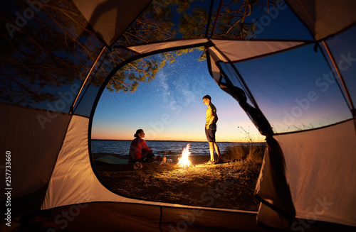 View from inside tourist tent at sunset. Young tourist couple, man and woman resting at bonfire, preparing food on gas burner on sea shore on evening starry sky and crystal sea water background.