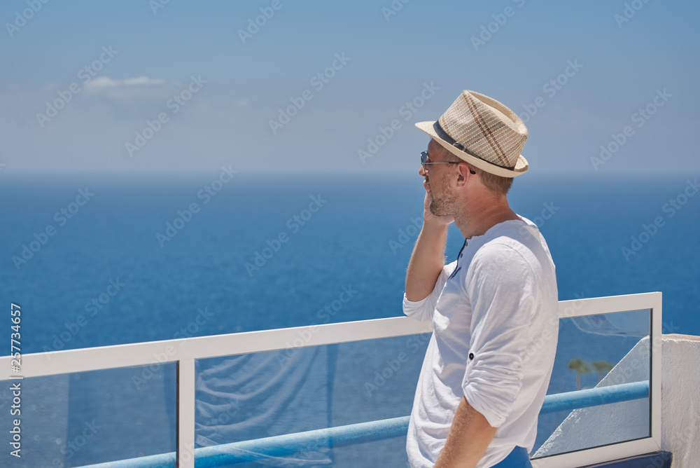 European man in a sunhat and sunglasses is talking on mobile phone, while having a rest at the beautiful resort. He is sharing his emotions.