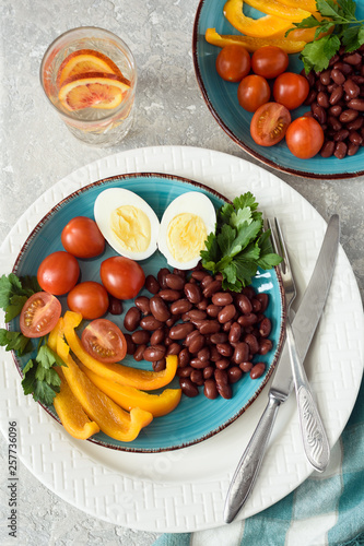 Healthy food, red beans, tomatoes, boiled egg, pepper with parsley in a plate with devices and a glass of water with orange on a gray background top view.