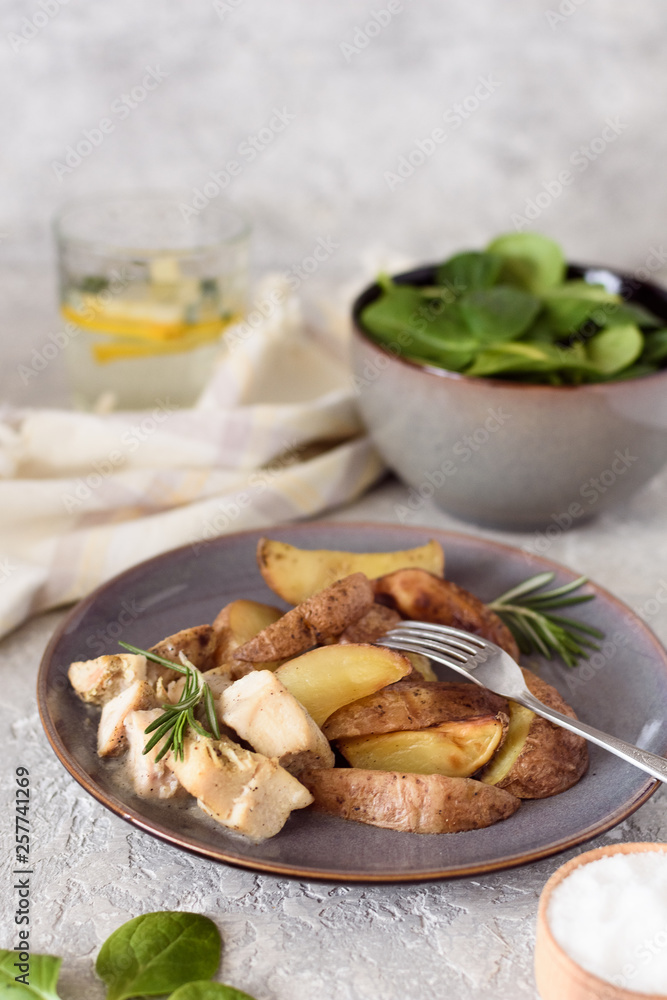 Baked potatoes sliced ​​with chicken fillet in a creamy sauce in a plate on the background of a salad bowl with spinach and a glass of water and lemon.