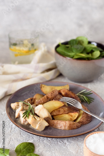 Baked potatoes sliced ​​with chicken fillet in a creamy sauce in a plate on the background of a salad bowl with spinach and a glass of water and lemon.