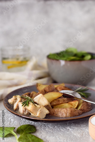 Baked potatoes sliced ​​with chicken fillet in a creamy sauce in a plate with a fork on the background of a salad bowl with spinach and a glass of water and lemon.