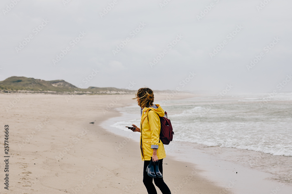 Young woman wearing yellow rain jacket strolling along North Sea Shore barefoot carrying her shoes in the hand Young woman wearing yellow rain jacket strolling along North Sea Shore barefoot carrying her shoes in the hand
