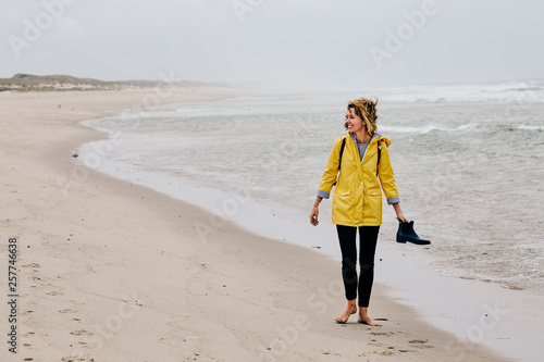 Young woman wearing yellow rain jacket strolling along North Sea Shore barefoot carrying her shoes in the hand photo