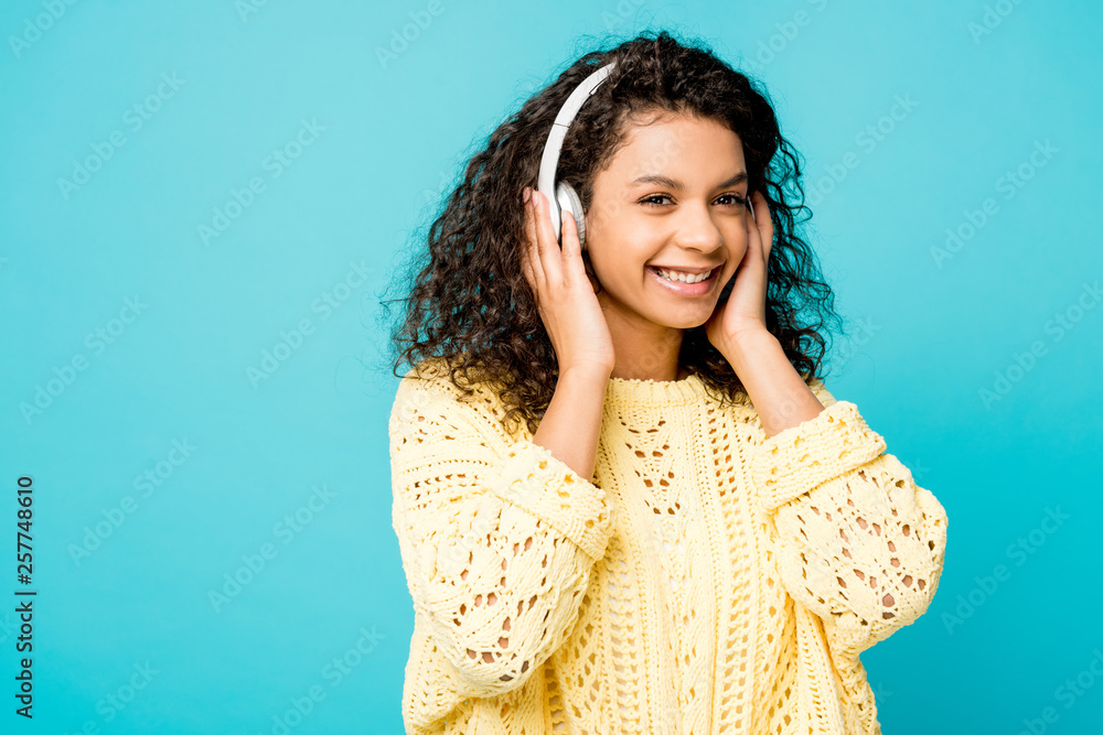 Fototapeta premium happy curly african american girl listening music in headphones isolated on blue