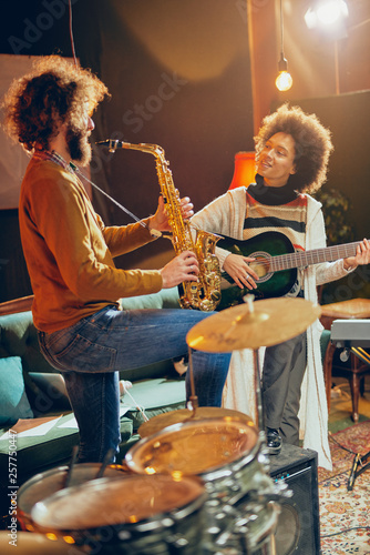 Mixed race woman playing acoustic guitar while man playing saxophone. Home studio interior.