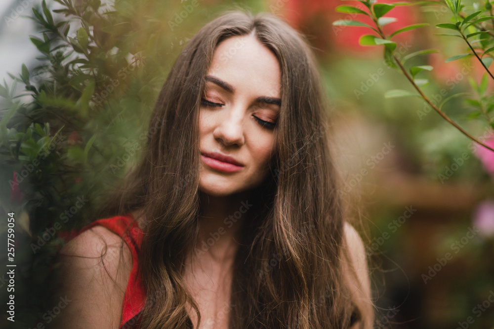 Naklejka premium Young beautiful caucasian woman in glass greenhouse among colorful azalea flowers. Art portrait of a long-haired girl wearing a romantic red dress.