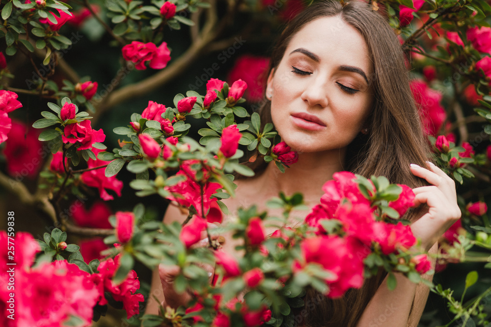 Young beautiful caucasian woman in glass greenhouse among colorful azalea flowers. Art portrait of a long-haired girl wearing a romantic red dress. Young beautiful caucasian woman in glass greenhouse among colorful azalea flowers. Art portrait of a long-haired girl wearing a romantic red dress.
