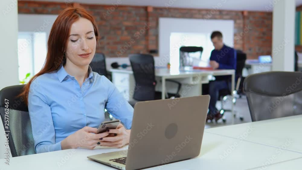 young adult employee sitting at the white desk in open space office startup company. redhead business woman holding smartphone texting message or chatting online on the background co workers works young adult employee sitting at the white desk in open space office startup company. redhead business woman holding smartphone texting message or chatting online on the background co workers works