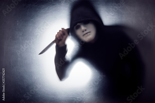 Man in a hood and white mask with knife behind a dusty scratched glass